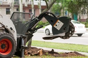 A skid steer loader with an auger attachment digging up dirt near a sidewalk.